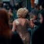 A performer in a sparkling gold dress moves through a crowded street, seen from behind while holding someone’s hand.