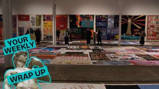 A large exhibition space filled with sections of the UK AIDS Memorial Quilt displayed across the floor and hanging on the walls, with visitors walking among them. A “Your Weekly Wrap Up” graphic appears in the lower-left corner.