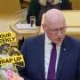 A bald man in glasses and a dark suit speaks in the Scottish Parliament chamber. A “Your Weekly Wrap Up” graphic with a classical statue and condom illustration appears in the lower-left corner.