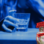 Close-up of a person holding a glass containing a clear drink under blue lighting. A “Your Weekly Wrap Up” graphic with a classical statue and condom illustration appears in the lower-right corner.